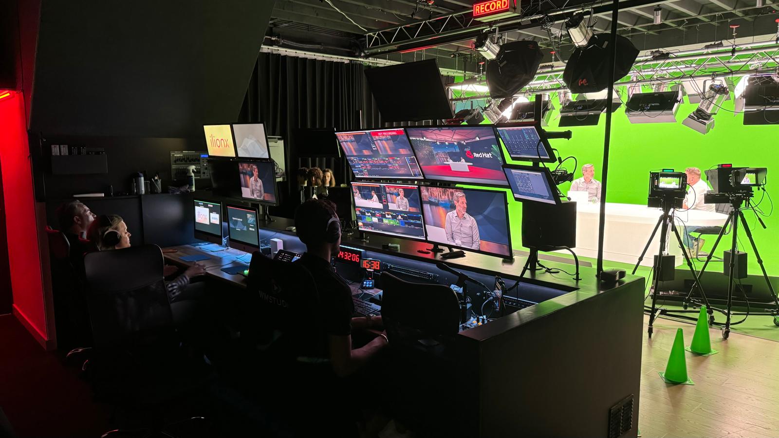 De regiekamer van de StoryYo-studio: twee mensen achter een wand met schermen, met daarnaast de set met camera's en een green screen.